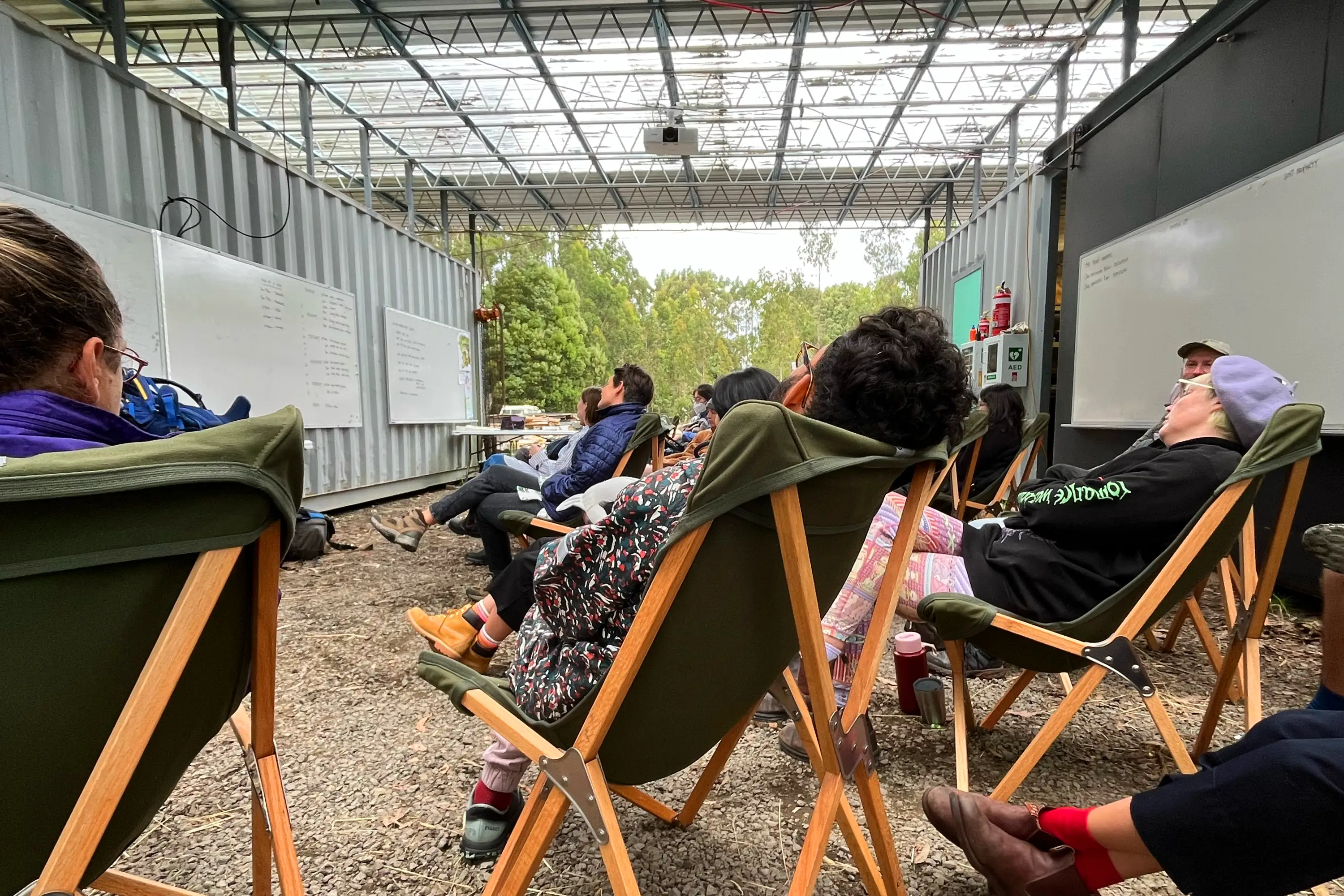 Under an exposed, metal truss roof, and between two shipping containers, a bunch of people sit in luxurious camping chairs on a gravel floor.