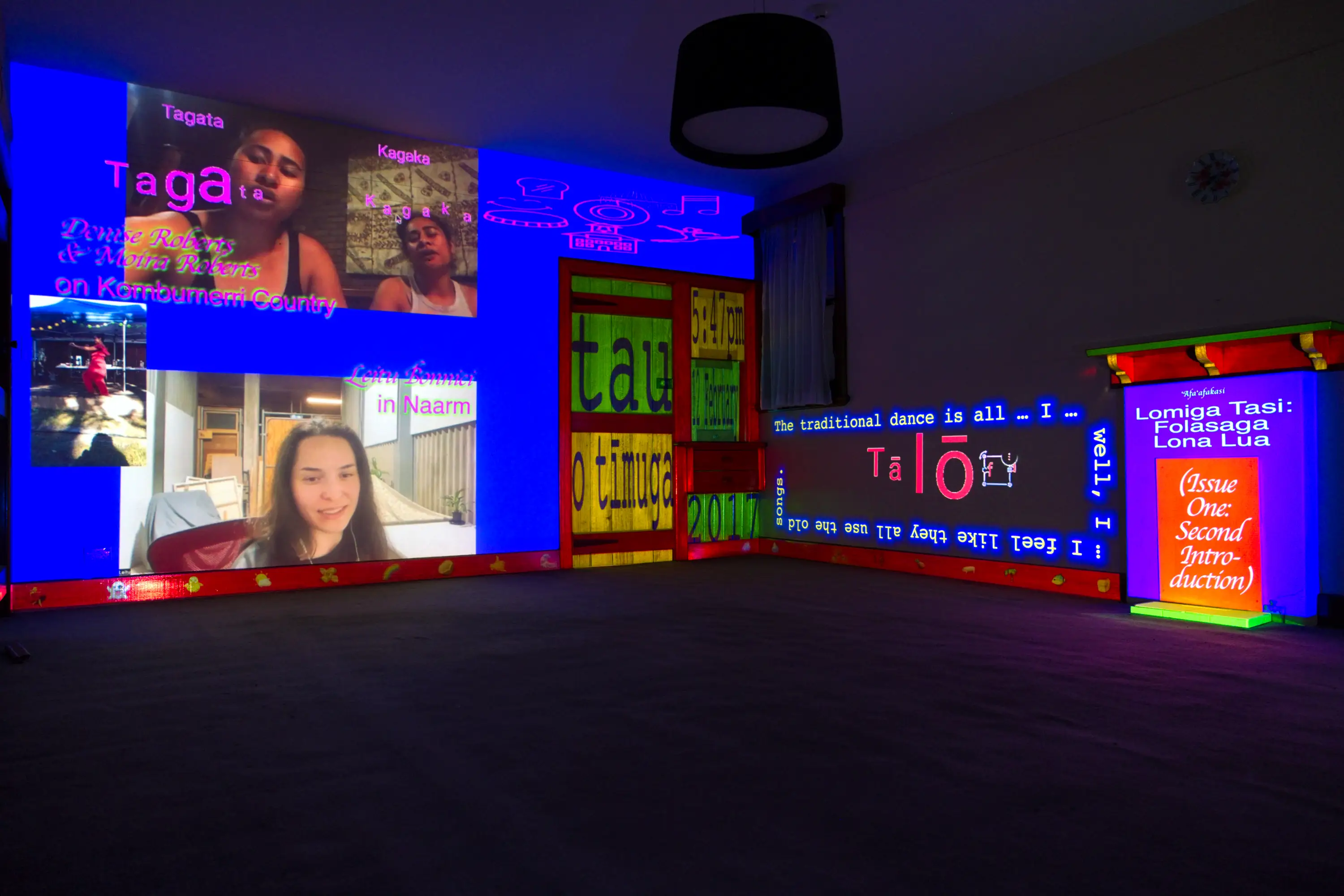 An overview of an installation in a large, empty, dark room. One wall is mostly covered in blue with Zoom footage, a phone video, writing and sketches over the top and the panels of the door are sectioned by colours and typography. The fireplace on the perpendicular wall is multicoloured and partially framing video captions on the wall.