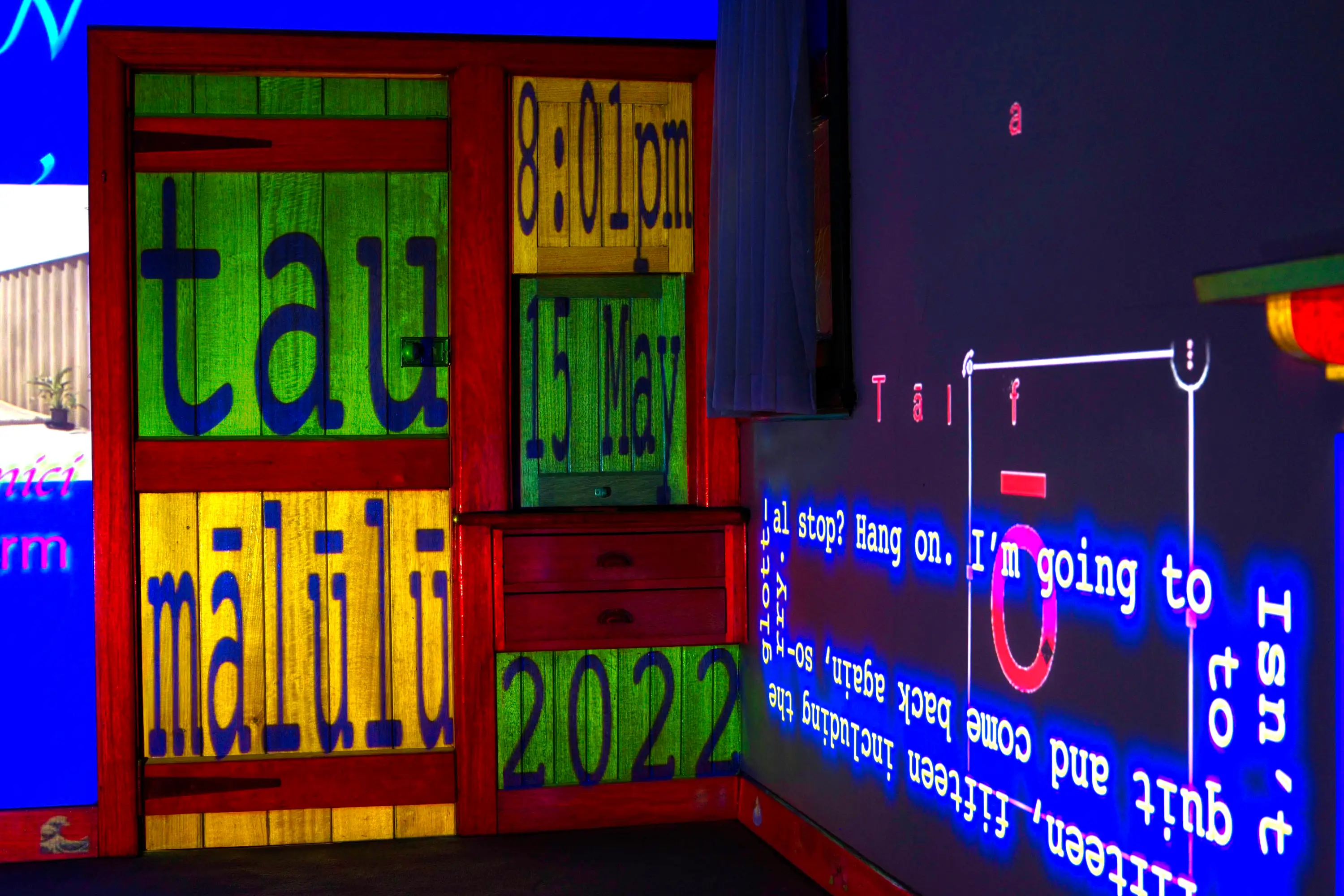 A close up of the coloured panels of the door with text that reads 'tau mālūlū', '8:01pm', '15 May', and '2022'. On the perpendicular wall is a video caption that spirals inward and is yellow with a blue glow, intersecting with the fractured word 'tālofa' in red.