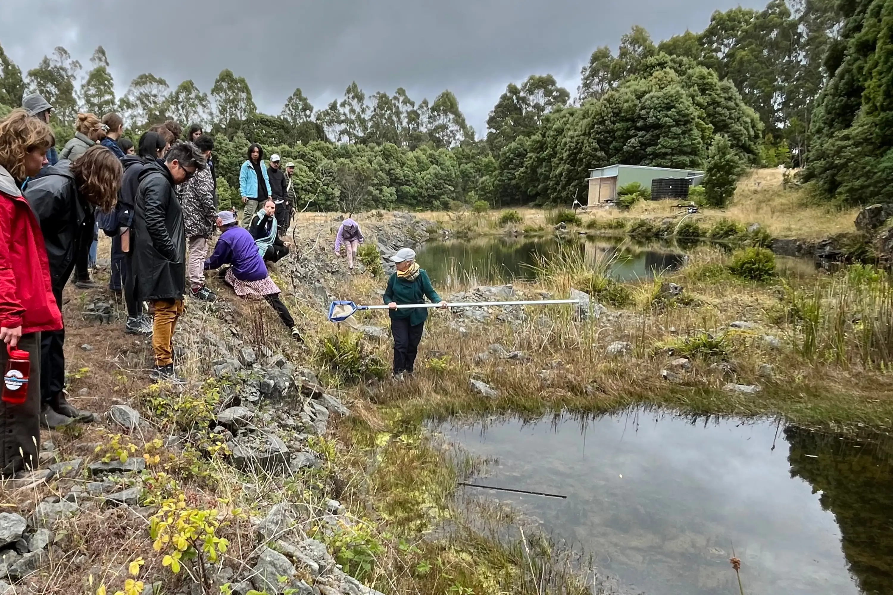 A group of people stand beside a large pond in the middle of a quarry, with one person holding a net with a very long handle.